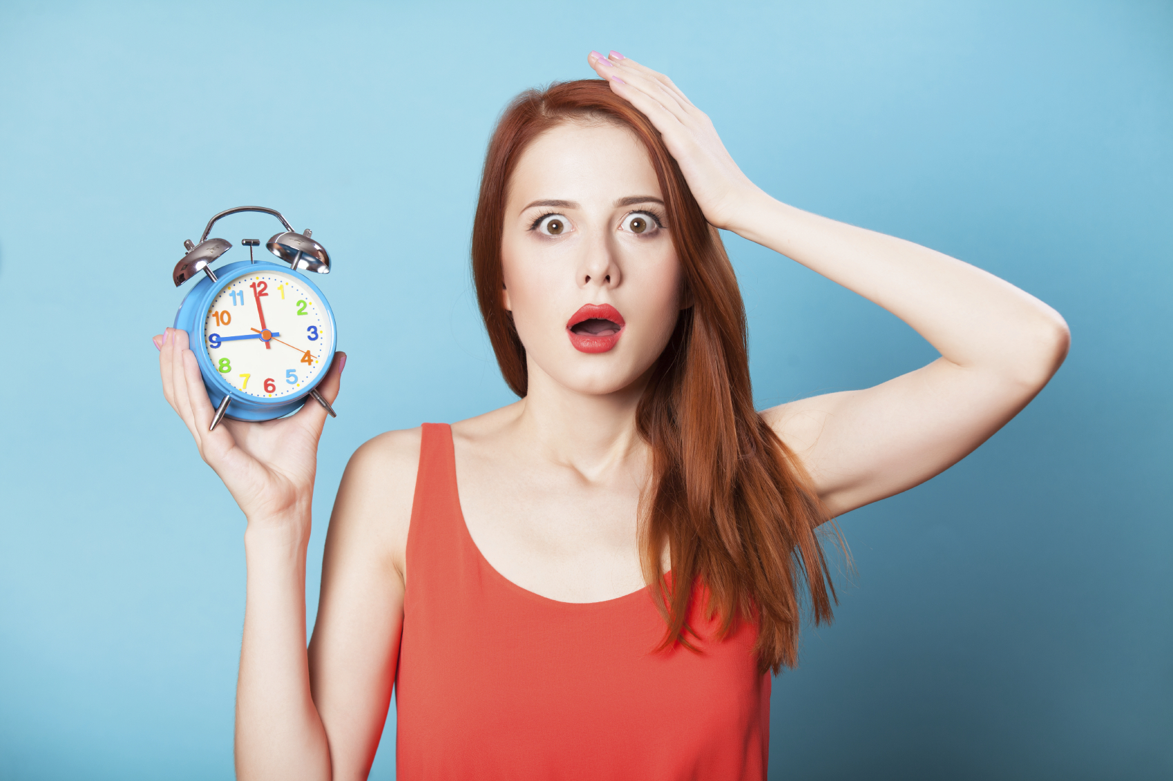 Surprised redhead women woth alarm clock on blue background.